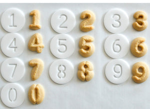 A set of 10 white plastic cookie press disks with numbers 0 through 9 imprinted on them, displayed on a light background.